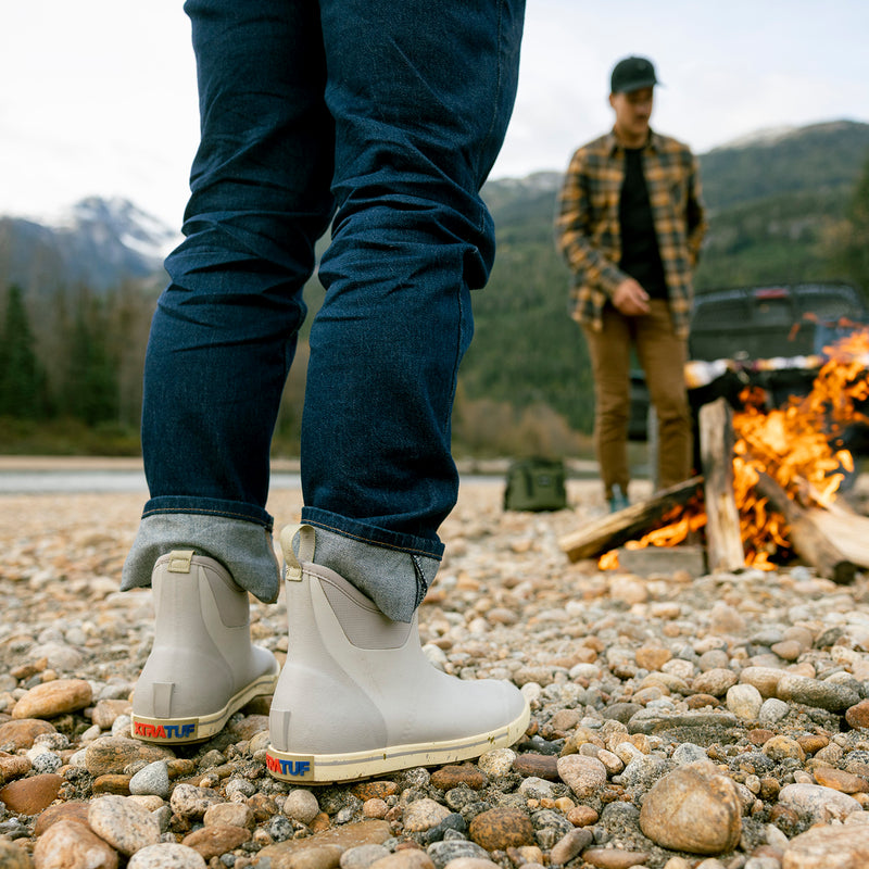 Person wearing gray boots on a rocky ground with another person and a campfire in the background.