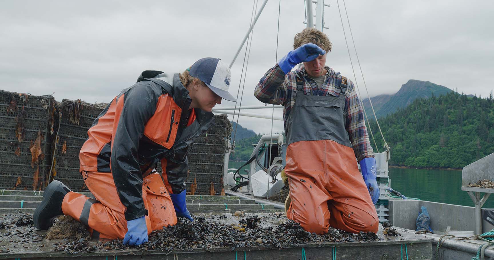 Two people in orange rain gear and blue gloves working on a boat with mountains in the background