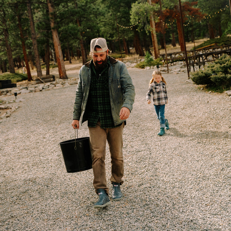 Man walking with a child in a forested area, holding a black bucket.
