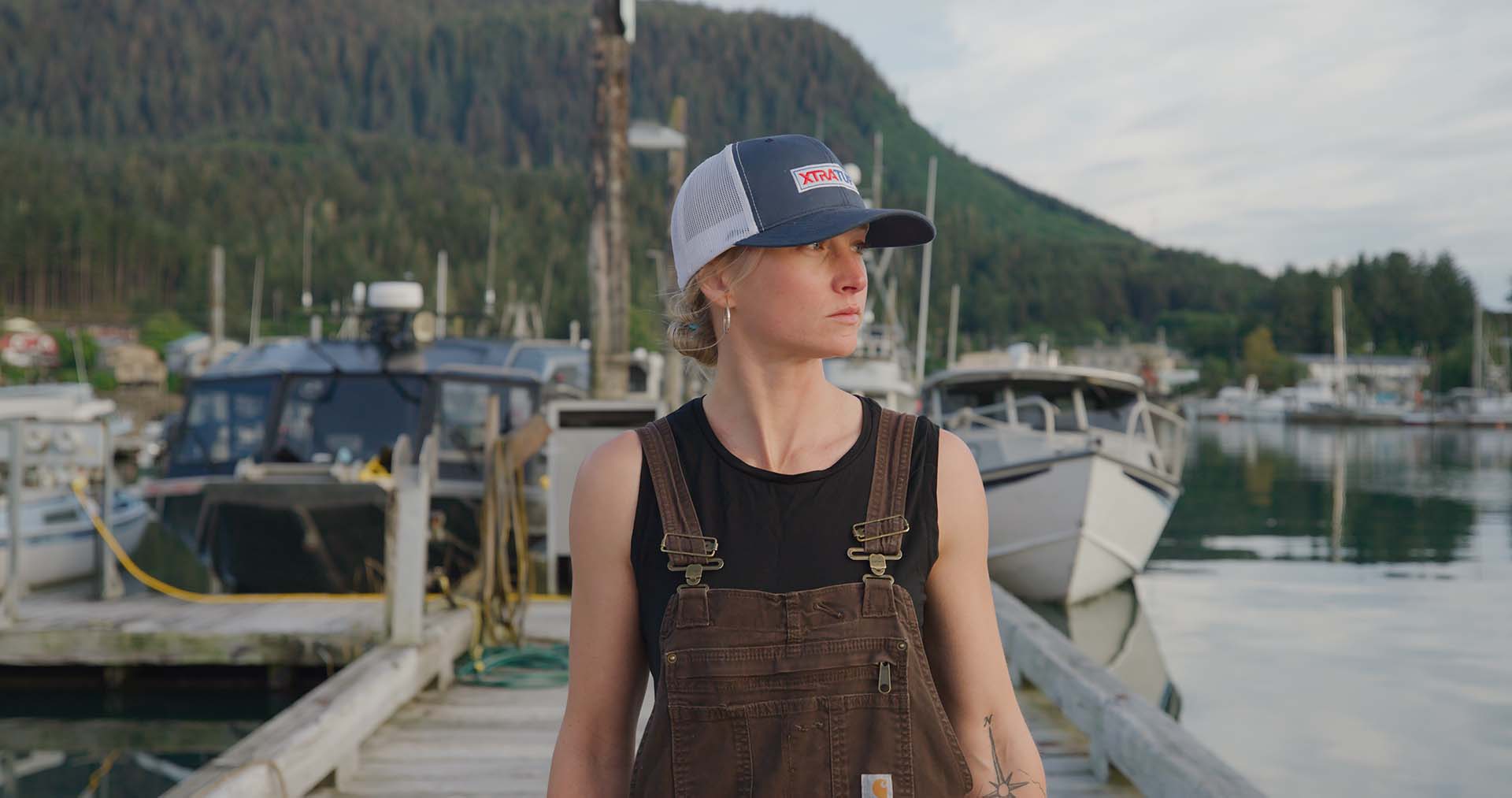 Person wearing a cap and overalls standing by a dock with boats and mountains in the background