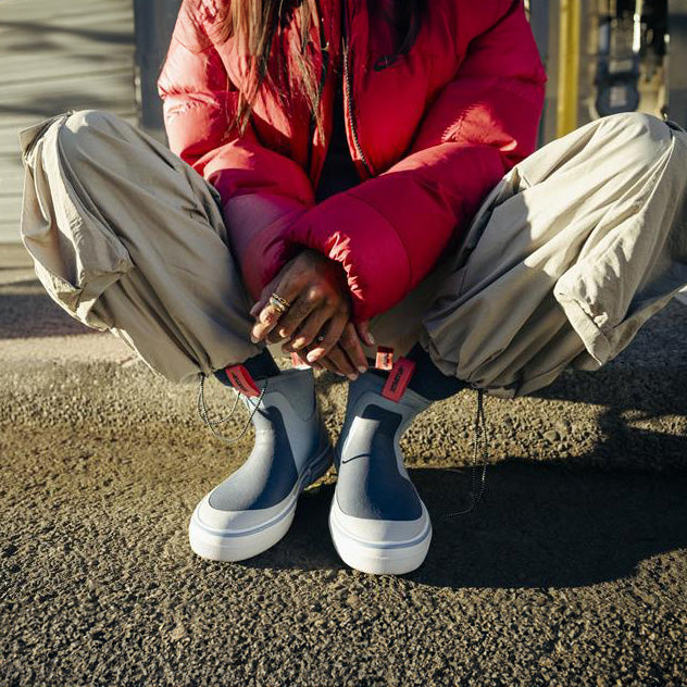 Person wearing red jacket and beige pants sitting on the ground with gray shoes.