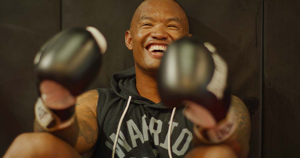 Man wearing boxing gloves and a black hoodie with 'Napoli' text, smiling.