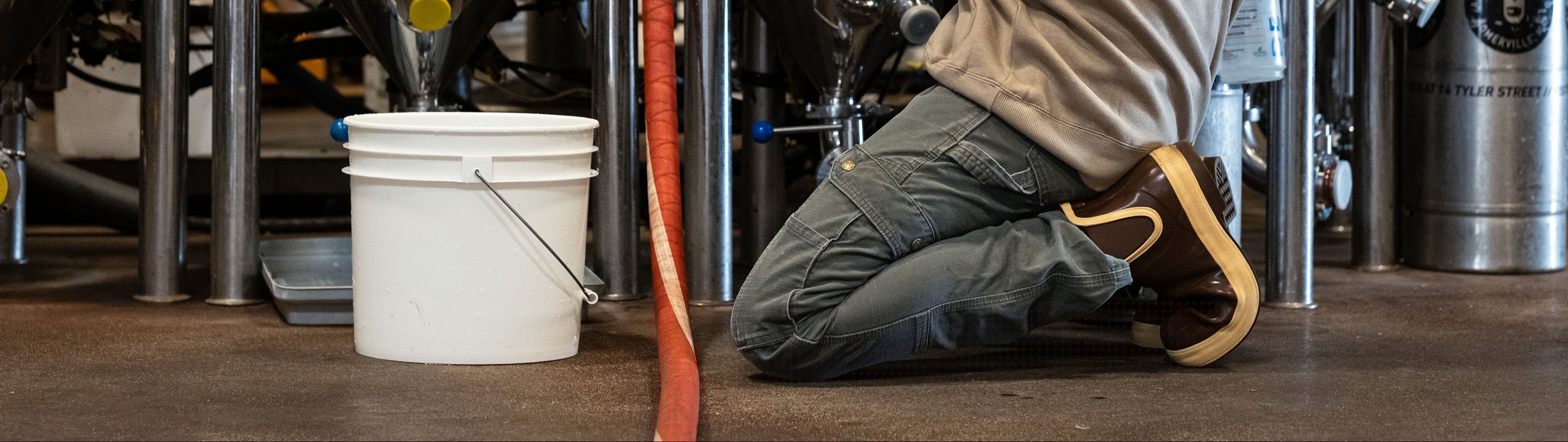 Person working in a brewery setting with large metal tanks and equipment.