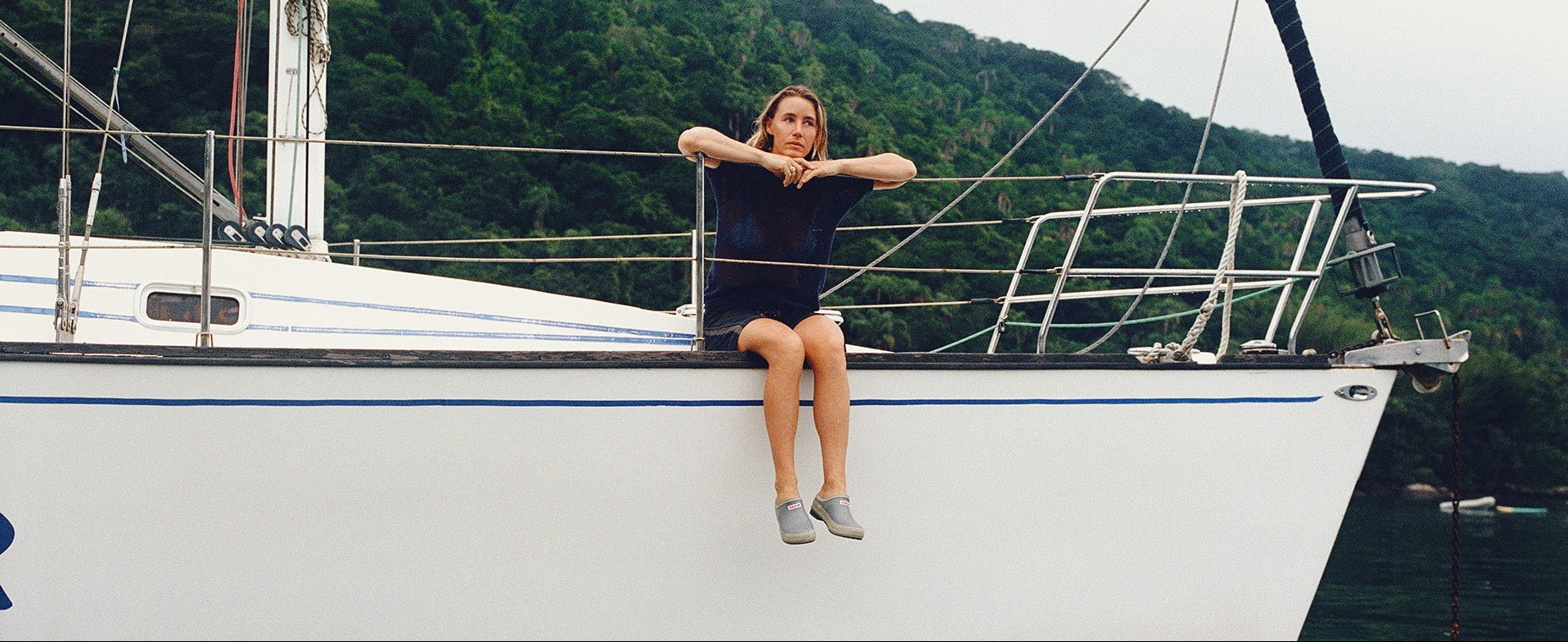 A woman sits on the side of a white sailboat moored in calm water, with a green forested hill in the background under an overcast sky.