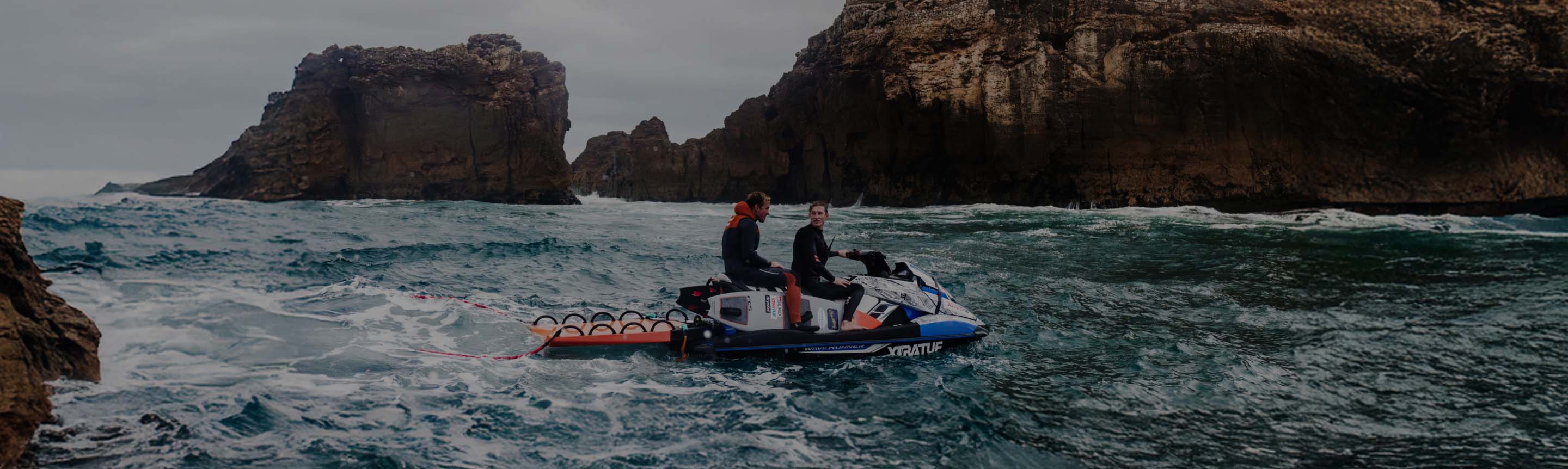 Two people on a jet ski in rough ocean waters with rocky cliffs in the background