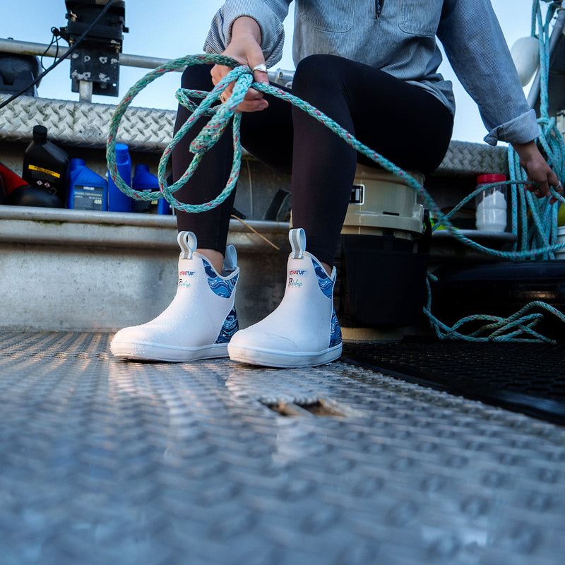 Person wearing white boots with blue patterns on a boat deck, holding green rope.