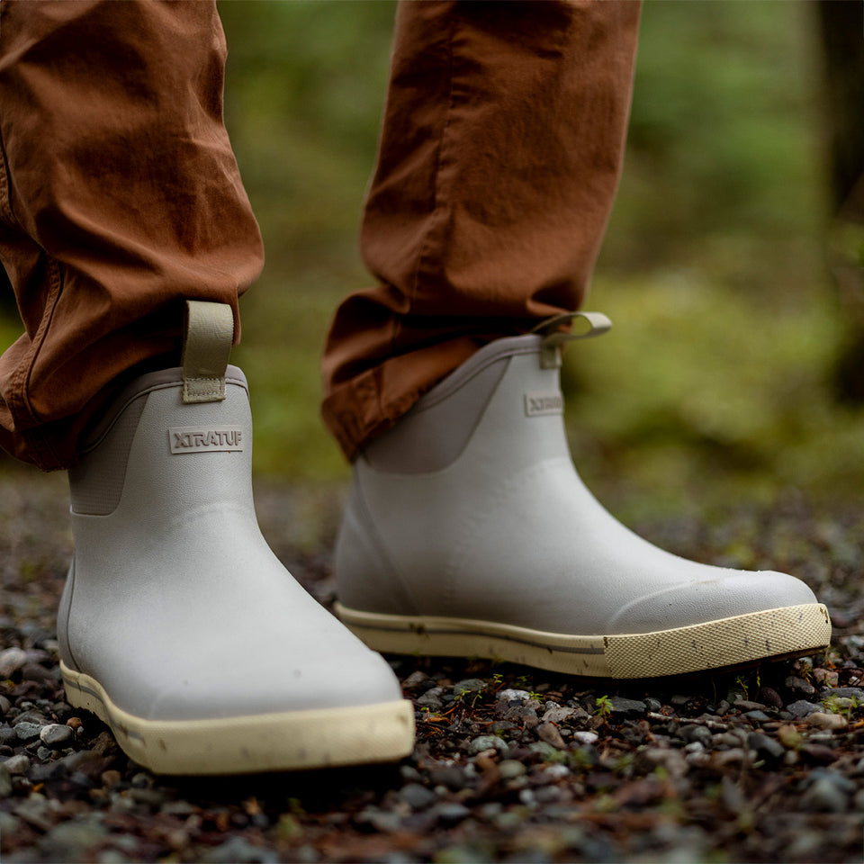 White rubber boots with beige soles worn by a person in brown pants on a natural ground.