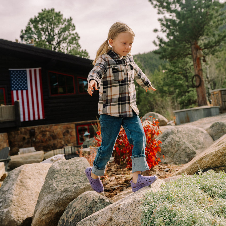 Child playing on rocks in a natural setting with a cabin and American flag in the background
