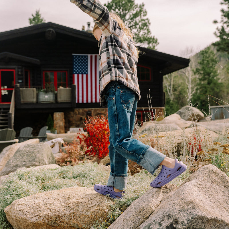 Person in plaid shirt and jeans jumping on rocks with a cabin and American flag in the background