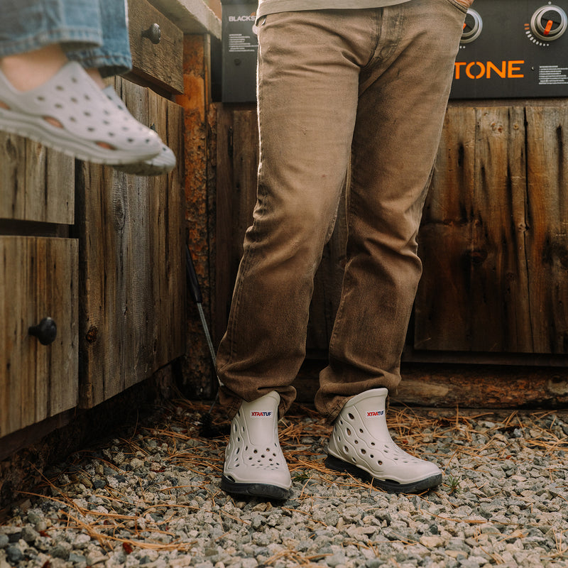 Person wearing brown pants and white Crocs shoes standing on a gravel surface with a wooden background.