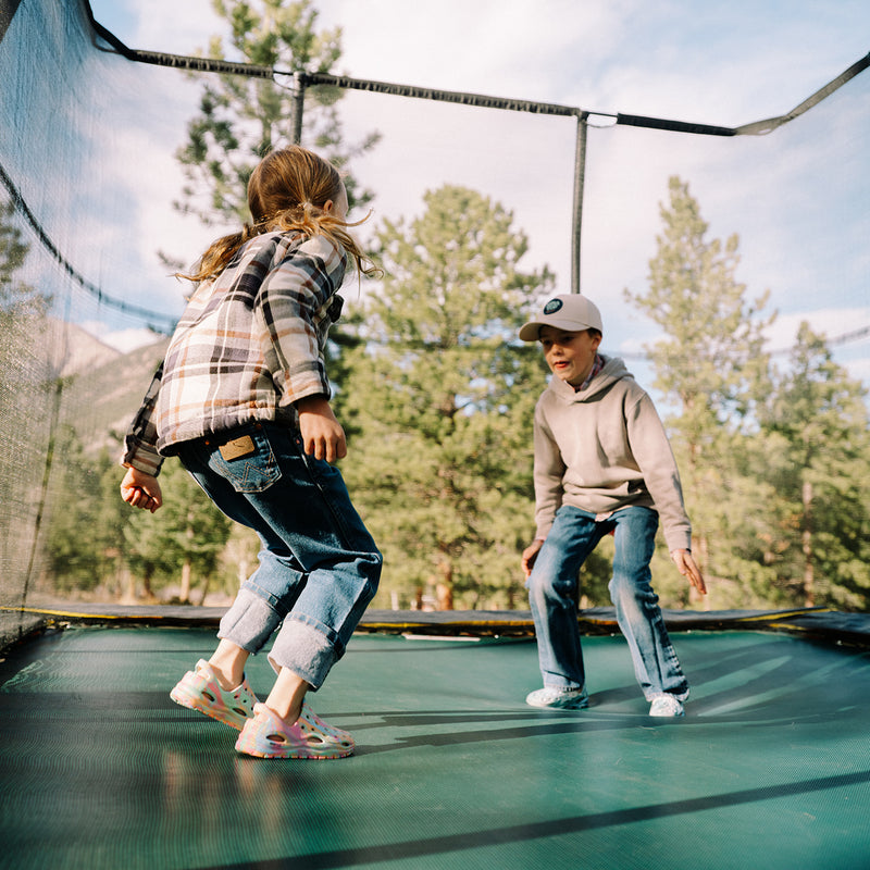 Two children playing on a trampoline outdoors with trees in the background