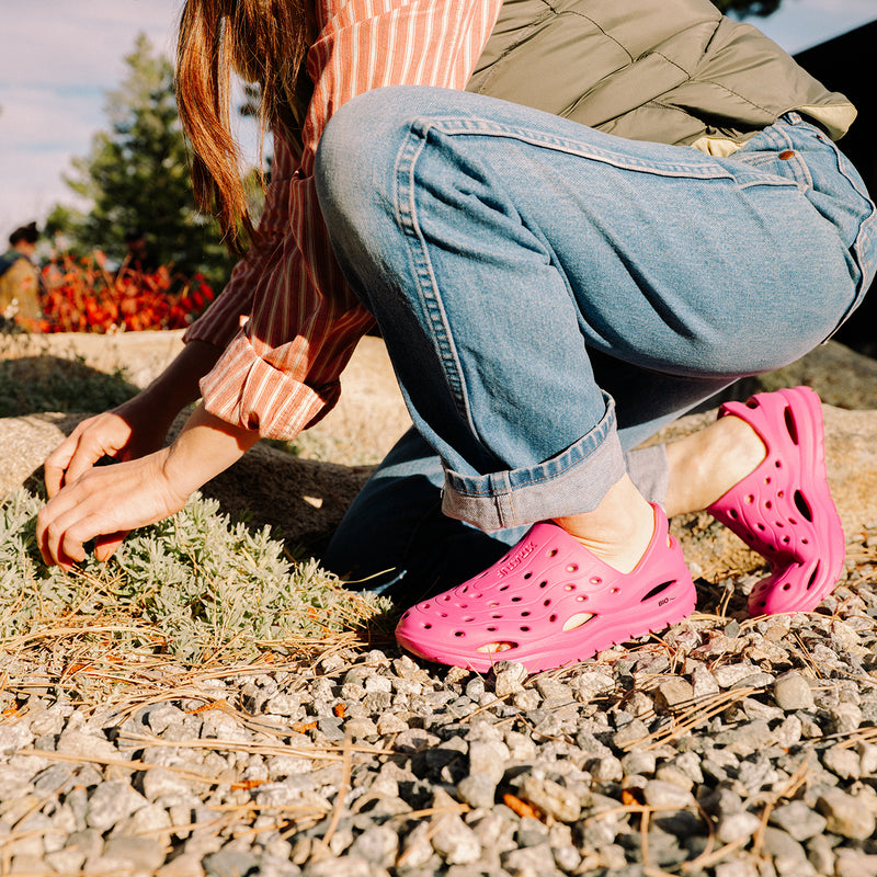 Person wearing pink clogs and blue jeans sitting on a stone path outdoors.