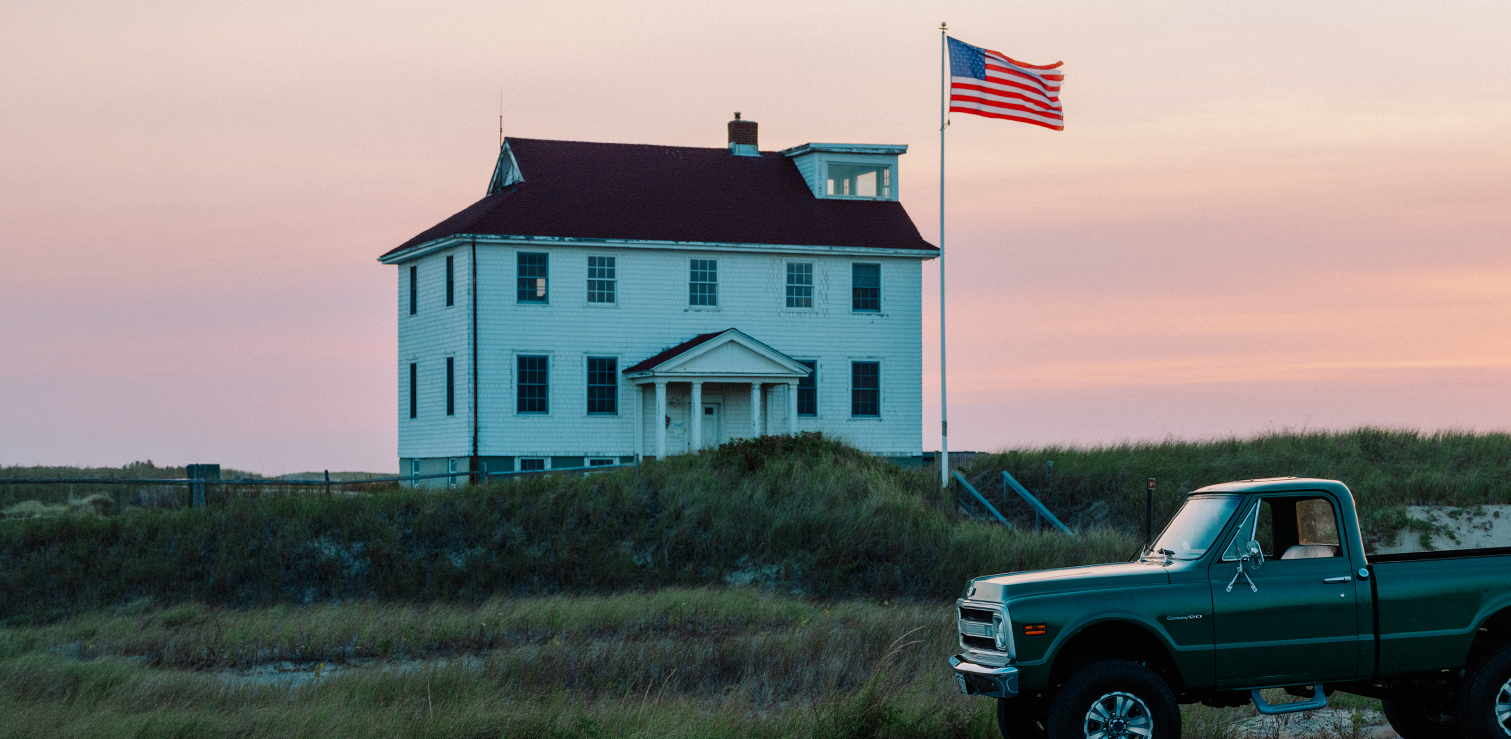 White house with an American flag on a grassy area, with a green truck parked nearby.
