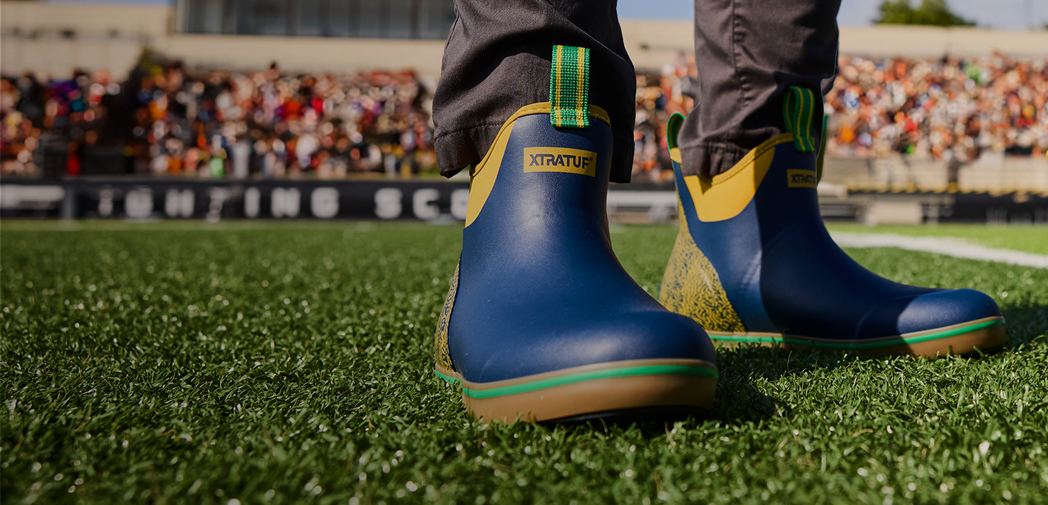 A close-up of a person's legs standing on a green turf field, wearing blue rubber boots with yellow and green accents. The brand name 'XTRATUF' is visible on the front of the boots.